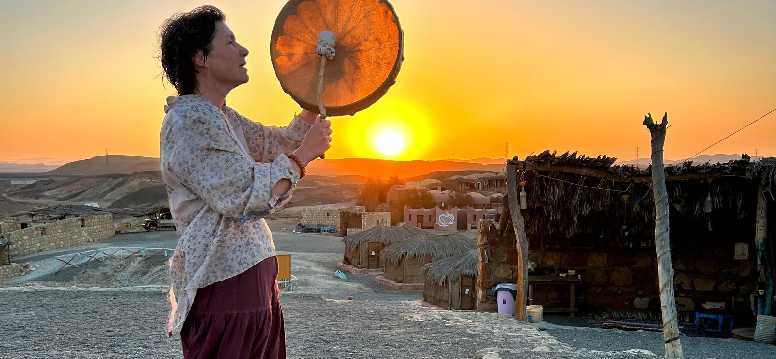 Nadiejda debout face à l'horizon, le tambour chamanique levé vers le soleil couchant — le soin vibratoire, entre ciel et terre
